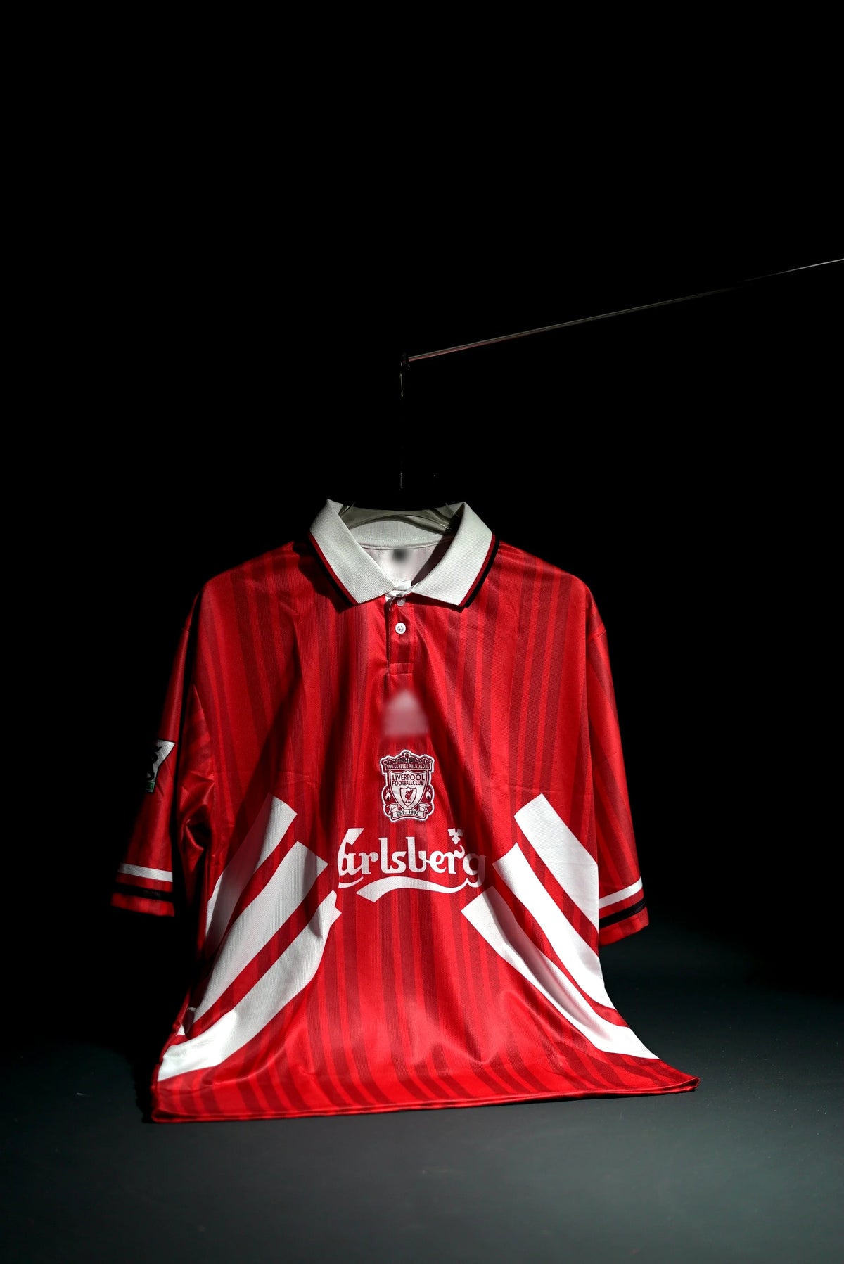 A vintage red and white striped Liverpool football jersey with a white collar, featuring the Carlsberg sponsor logo on the chest, displayed on a hanger against a dark background.