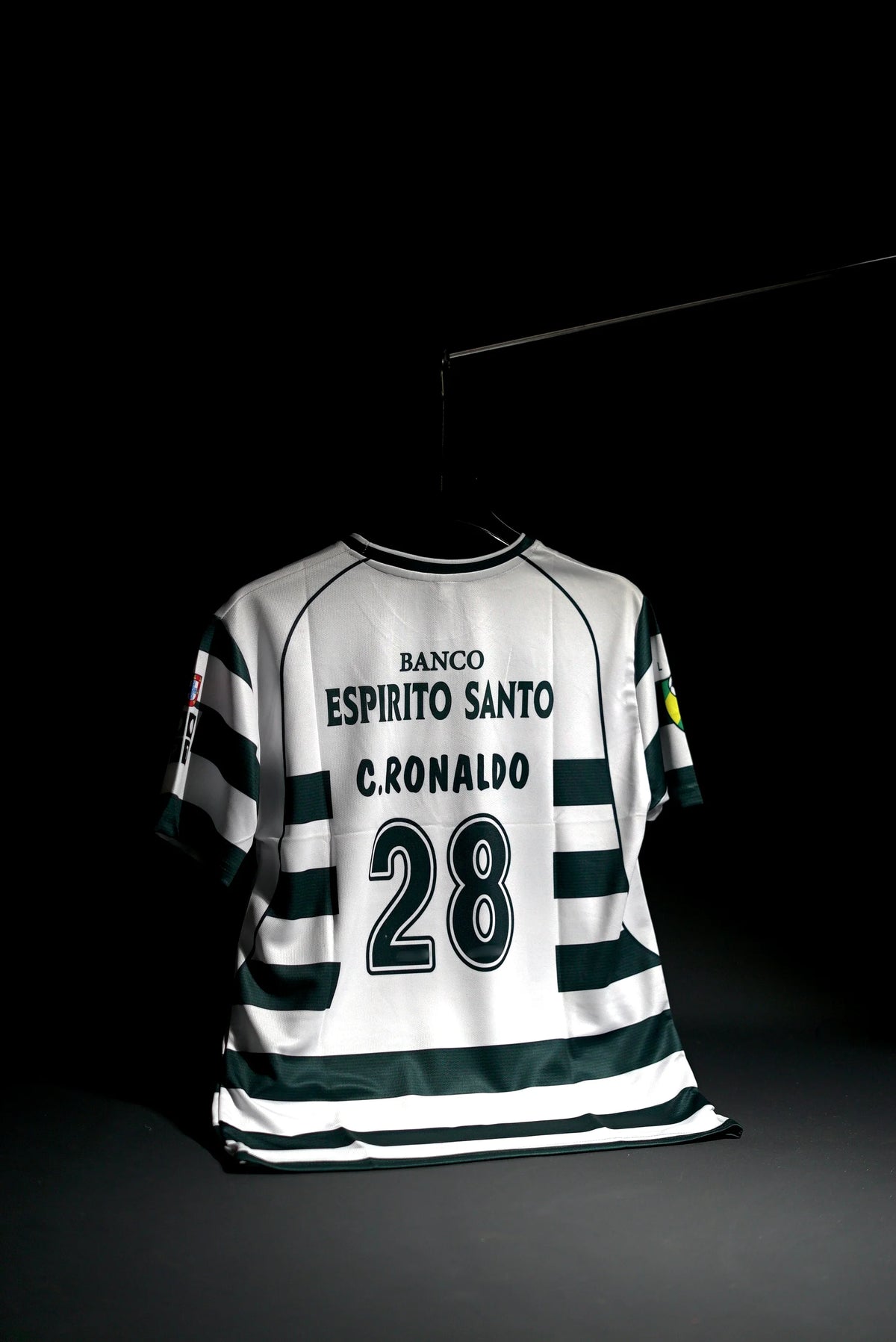 A white and dark green striped soccer jersey with 'BANCO ESPIRITO SANTO', 'C. RONALDO', and the number '28' printed on the back, hanging against a black background.